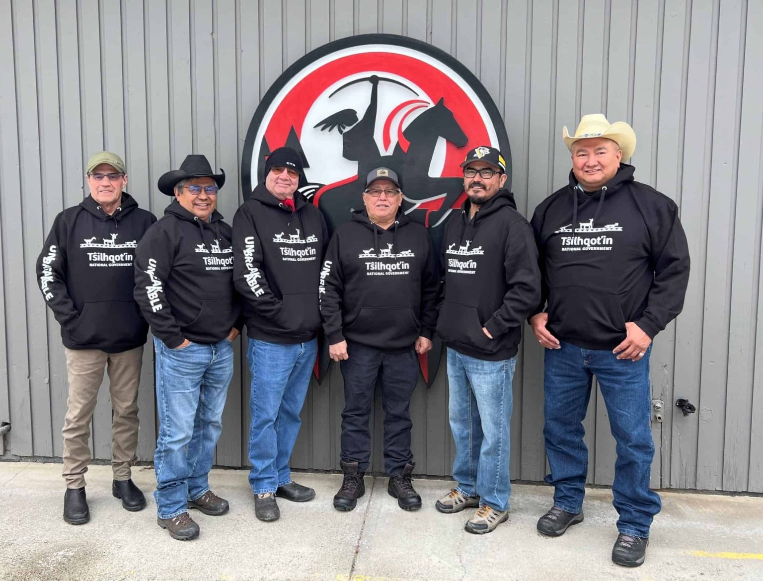 The six Tŝilhqot'in Nits'ilʔin-Qi (Chiefs) in 2026, standing before the Tŝilhqot'in National Government crest